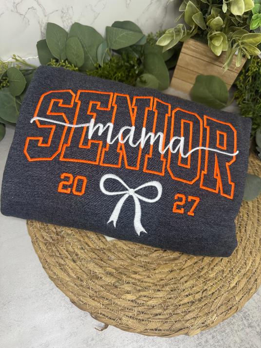 A folded dark heather gray sweatshirt with orange and white graphic text reading 'SENIOR mama 2027' and a bow design, displayed on a round woven mat with greenery in the background.