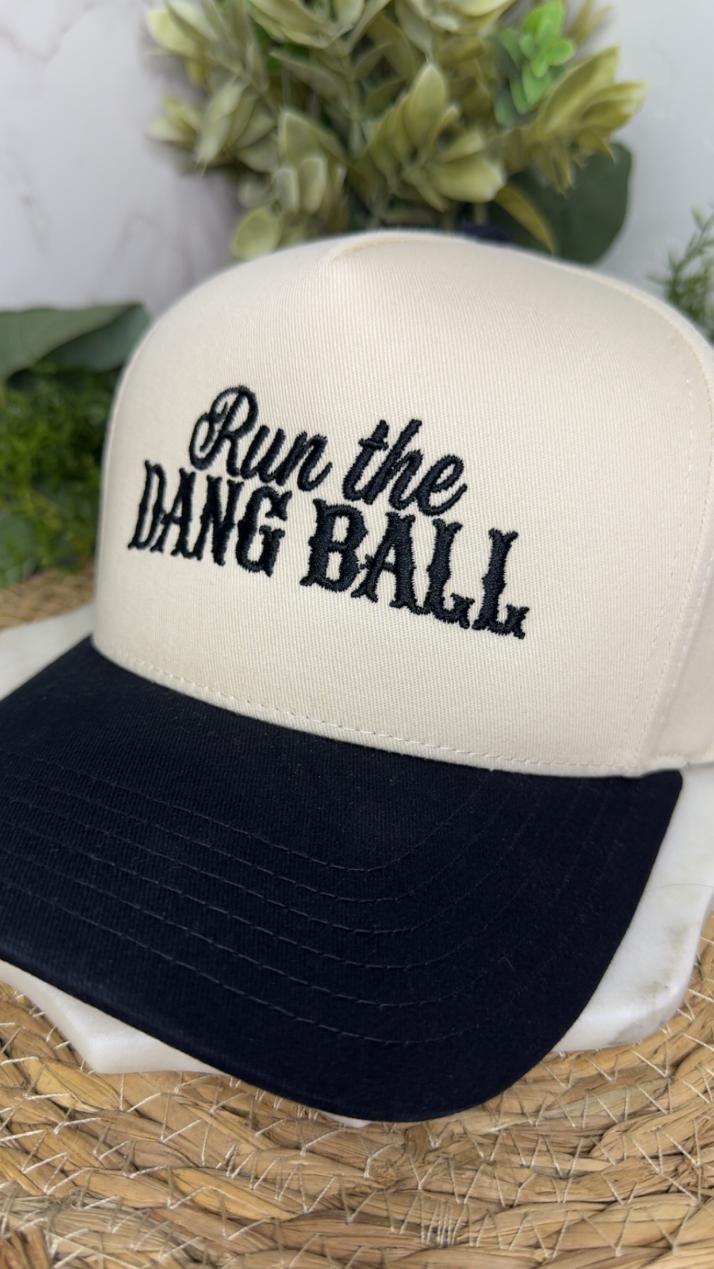 A cream and black trucker-style cap with the phrase 'Run the DANG BALL' embroidered in black script and block lettering on the front. The cap is displayed on a woven straw mat with blurred green foliage in the background.