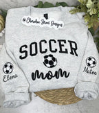 A light heather gray crewneck sweatshirt with 'SOCCER mom' printed in black on the front, featuring soccer ball graphics and personalized names 'Elena' and 'Mateo' on the sleeves. The sweatshirt is displayed flat on a woven mat with green foliage in the background.