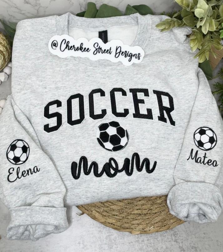 A light heather gray crewneck sweatshirt with 'SOCCER mom' printed in black on the front, featuring soccer ball graphics and personalized names 'Elena' and 'Mateo' on the sleeves. The sweatshirt is displayed flat on a woven mat with green foliage in the background.