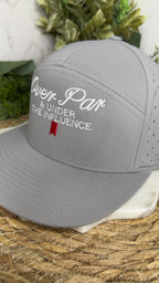 A light gray baseball cap with white embroidered text reading 'Oven Par & Under The Influence' and a small red ribbon accent, displayed on a woven mat with greenery in the background.