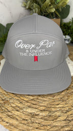 A gray snapback cap with white embroidered text reading 'Over Par & Under The Influence' and a small red flag detail on the front, displayed on a woven mat with greenery in the background.