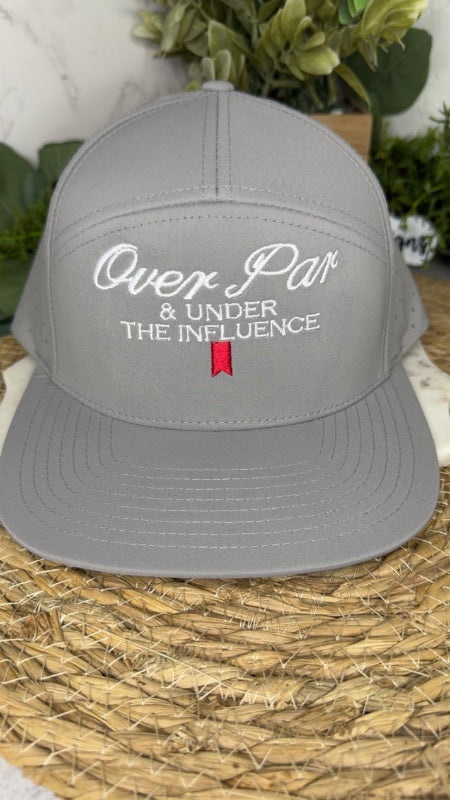 A gray snapback cap with white embroidered text reading 'Over Par & Under The Influence' and a small red flag detail on the front, displayed on a woven mat with greenery in the background.