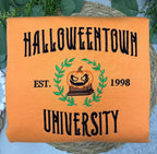 An orange folded fabric item, likely a sweatshirt or blanket, featuring embroidered text that reads 'HALLOWEENTOWN UNIVERSITY' with a central jack-o'-lantern inside a green wreath and 'EST. 1998'.