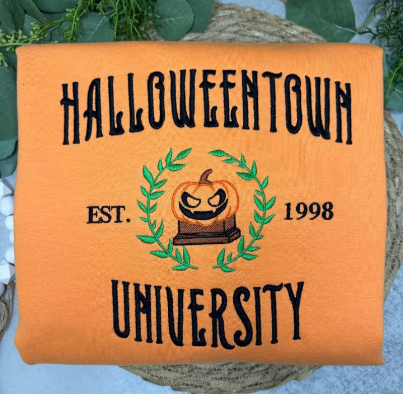 An orange folded fabric item, likely a sweatshirt or blanket, featuring embroidered text that reads 'HALLOWEENTOWN UNIVERSITY' with a central jack-o'-lantern inside a green wreath and 'EST. 1998'.